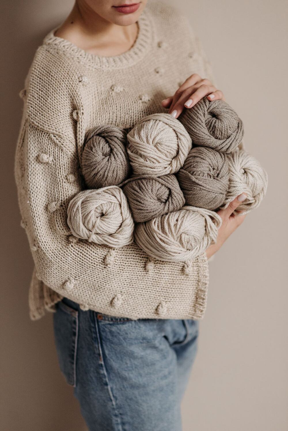 Person im beigen Strickpullover hält Wollknäuel in Creme- und Taupetönen